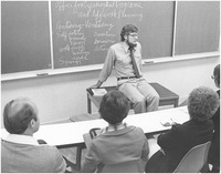 Ken Wood sitting on a table in a classroom with an outline of his Center-Venture model written on the chalkboard behind him .