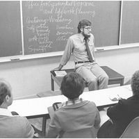 Ken Wood sitting on a table in a classroom with an outline of his Center-Venture model written on the chalkboard behind him .